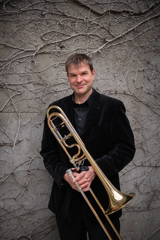 Jonathan Reycraft holds his trombone while wearing a dark suit and standing against a textured wall.