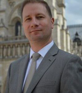 Johann Vexo, Organist, with a neutral expression as he stands in a light-colored suit and tie in front of Notre Dame Cathedral in Paris.